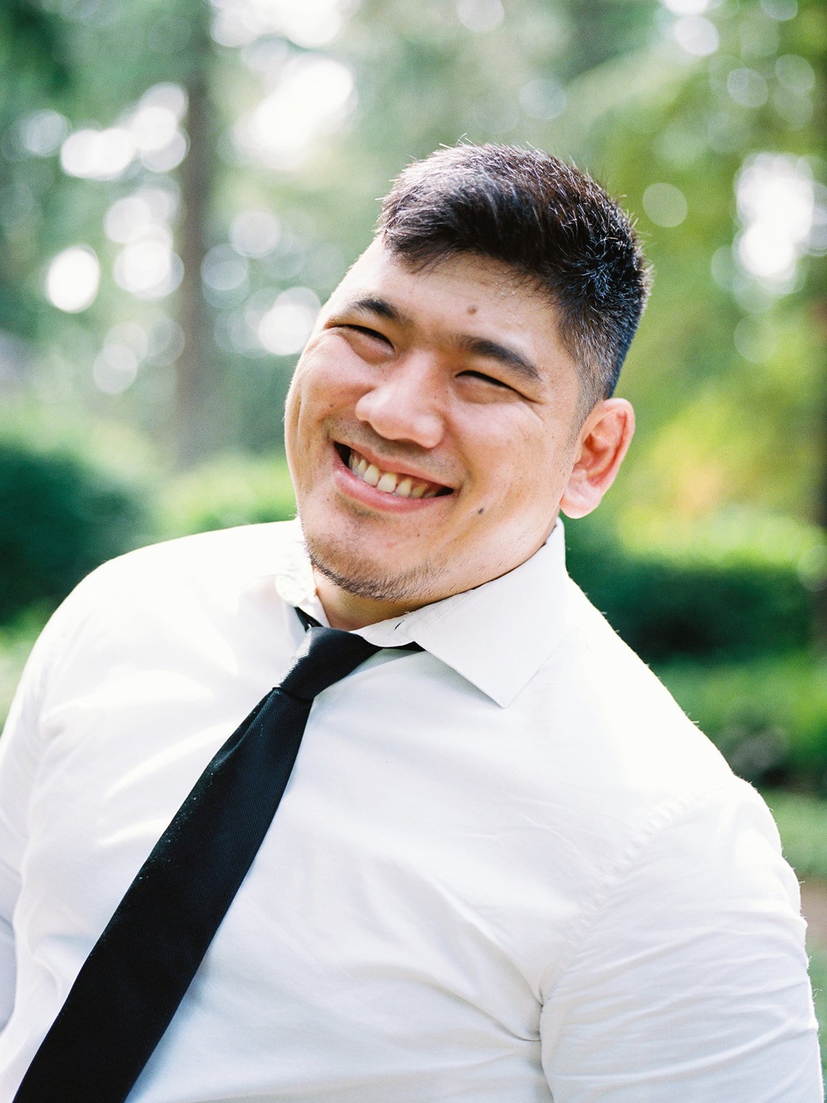 Close portrait of groom smiling outdoors in a white shirt and black tie with soft greenery behind him.