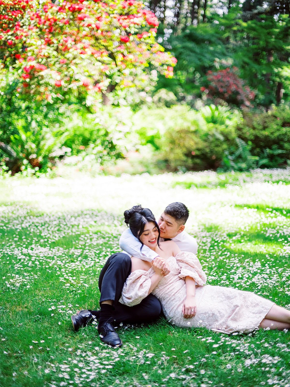 Couple sitting on grass covered in small white flowers beneath a blooming tree at The Grotto in Portland.