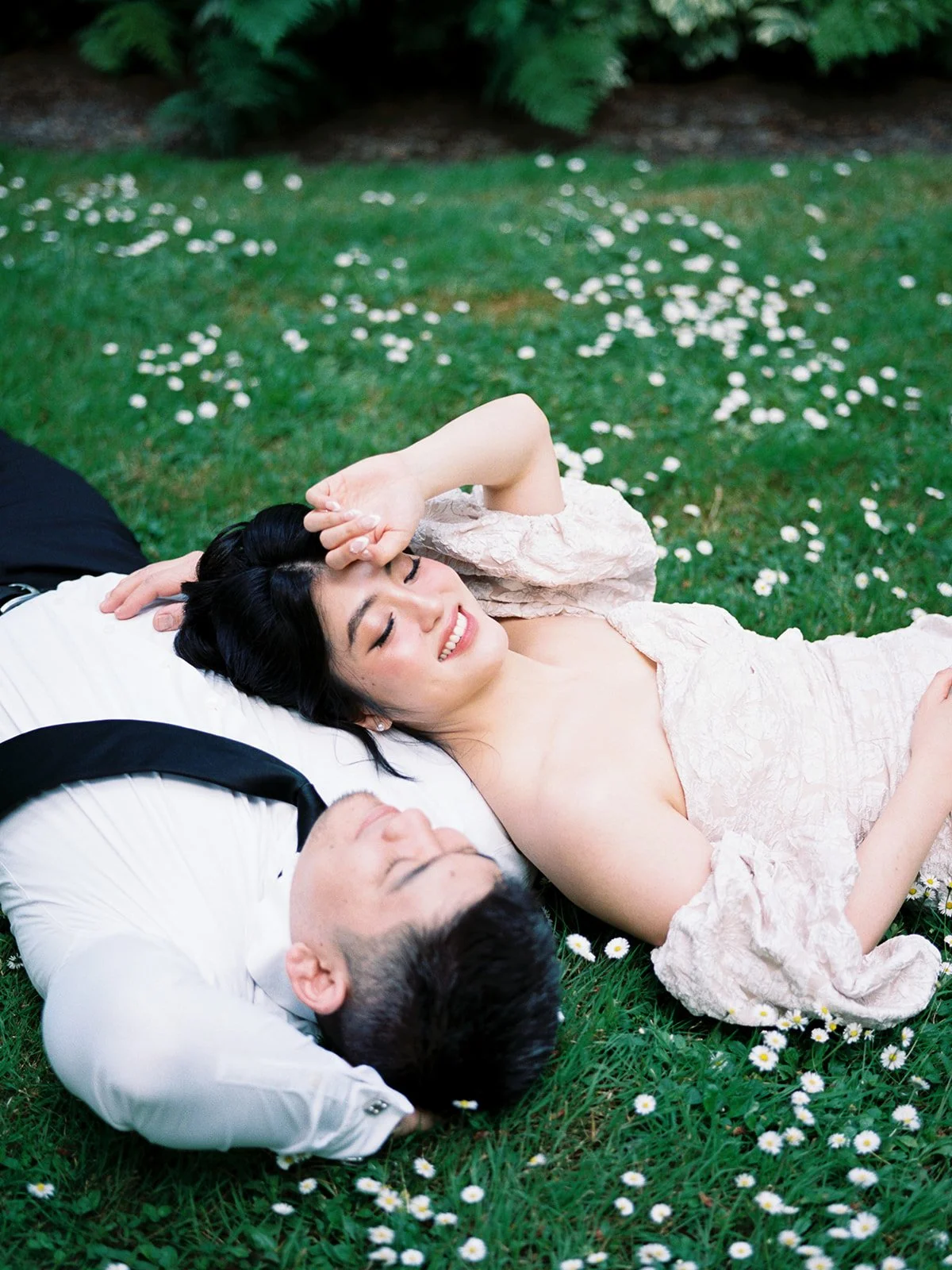 Couple lying on grass dotted with small white flowers, relaxing together in a garden setting.