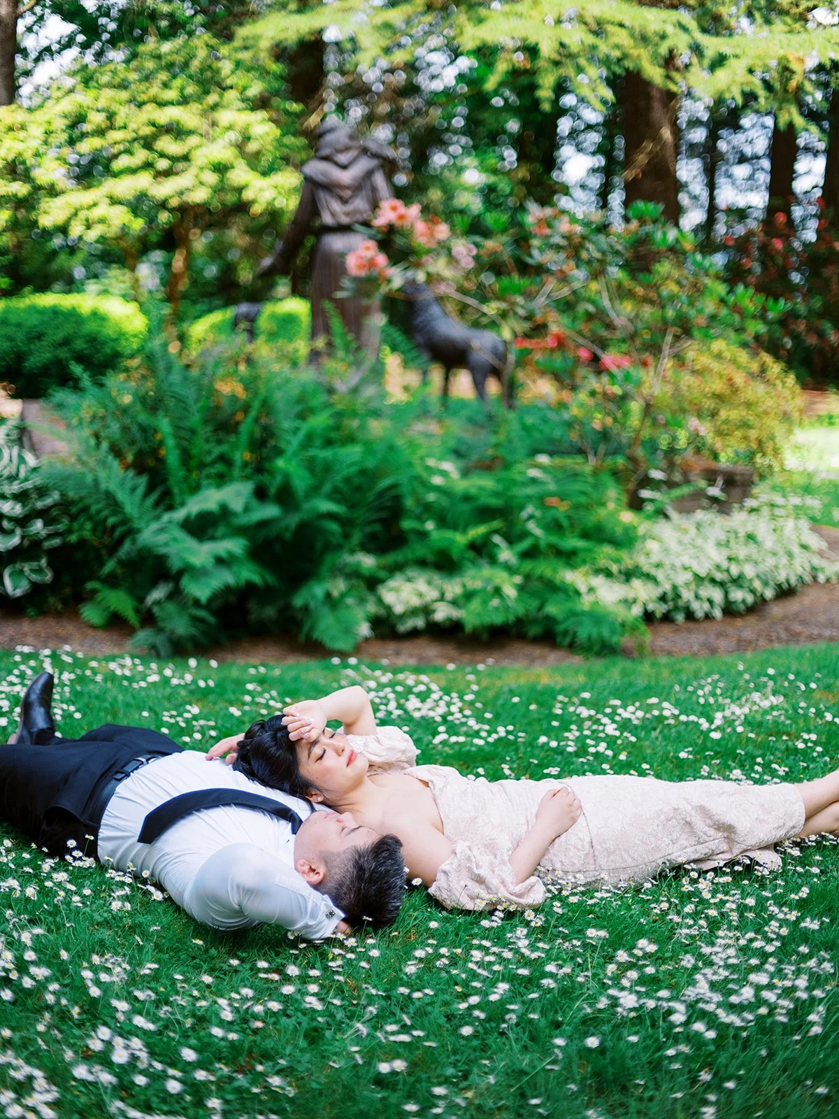 Wide view of couple lying in a grassy garden filled with small white flowers and surrounded by lush greenery.