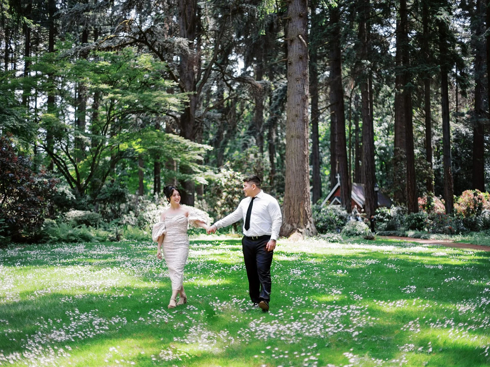 Couple holding hands while walking across a sunny lawn surrounded by tall trees and forest greenery.