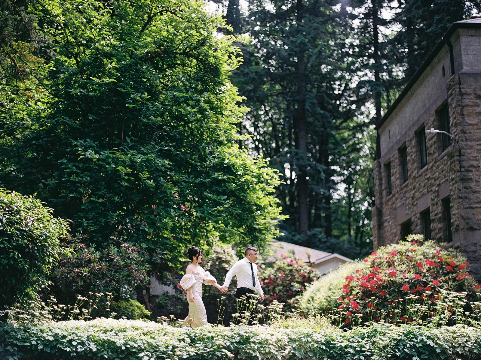 Couple walking hand in hand through a garden path beside stone buildings and blooming shrubs.