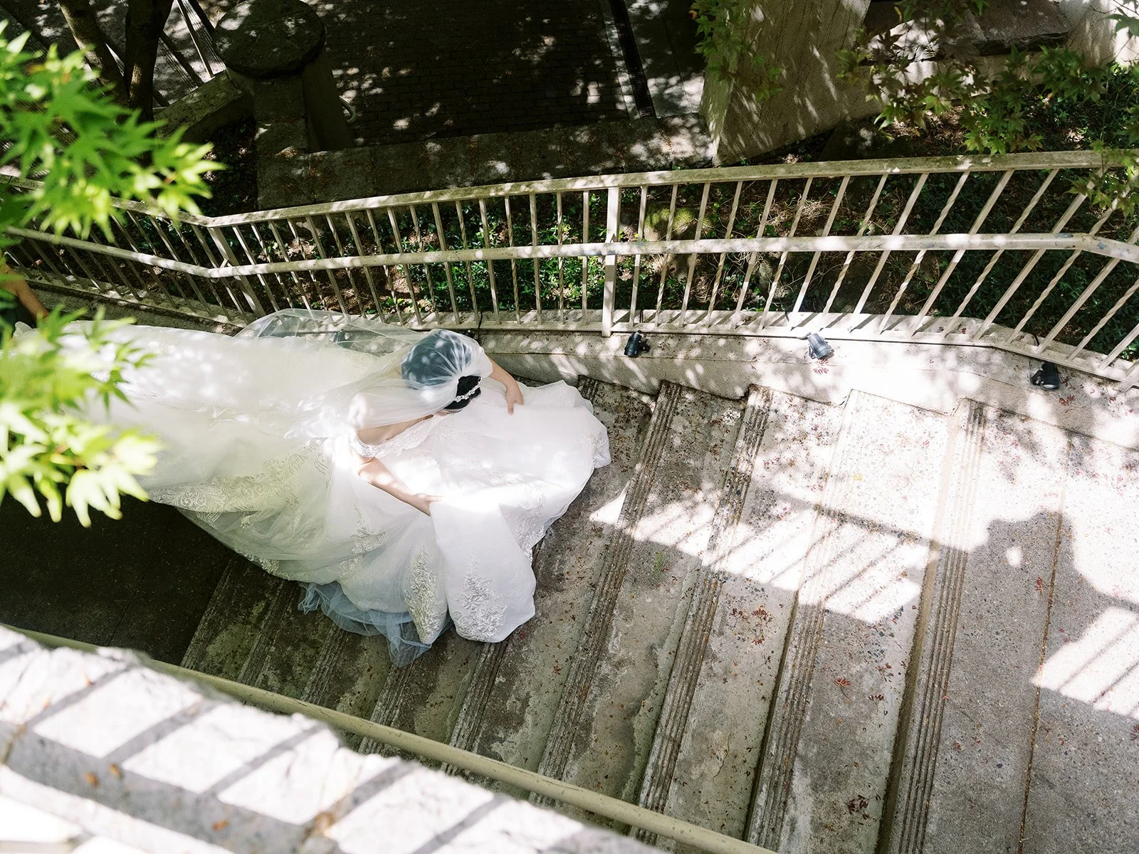 Bride in a flowing gown and veil walking down curved stone steps surrounded by trees and dappled sunlight.
