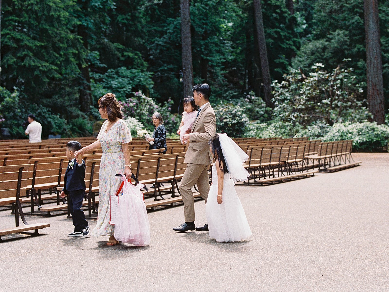 Family members walking through outdoor ceremony seating in a wooded garden setting before the wedding.