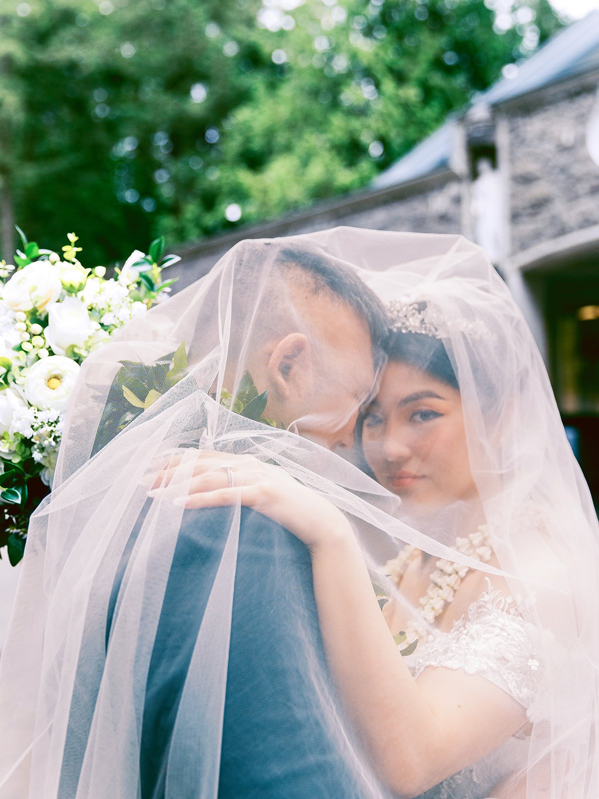 Bride and groom embracing beneath the bride’s veil with greenery and stone architecture behind them.