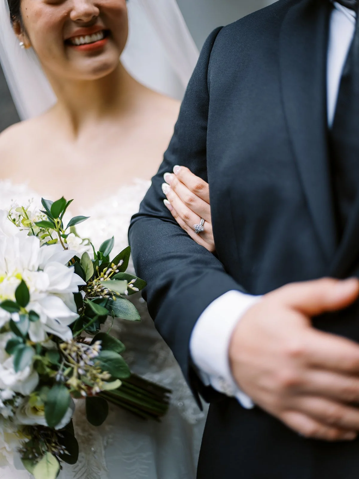 Close-up of bride’s hand with engagement ring resting on groom’s arm while holding a white wedding bouquet.