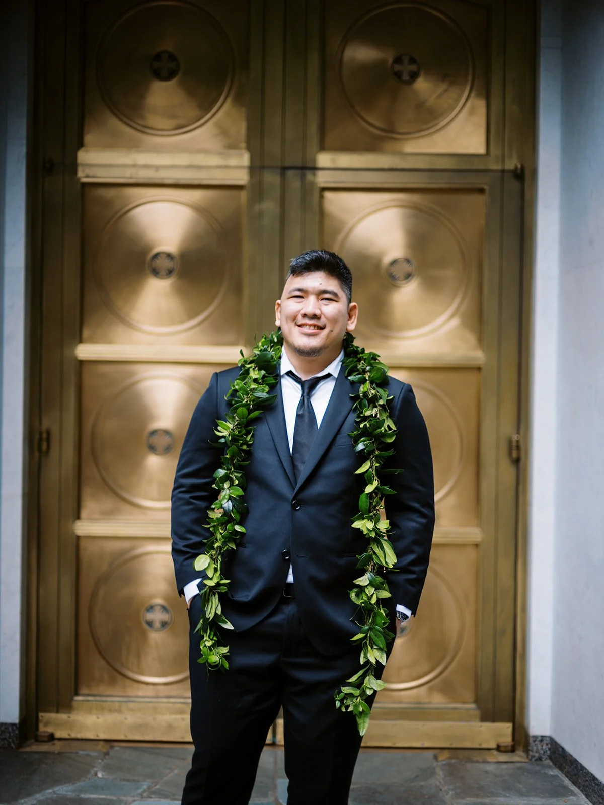 Groom wearing a green leafy lei standing in front of large golden church doors.