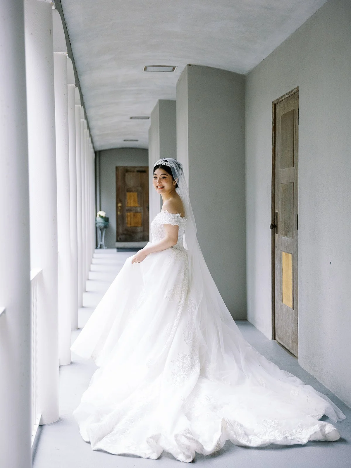 Bride smiling while standing in a bright arched hallway with her wedding gown and long veil.