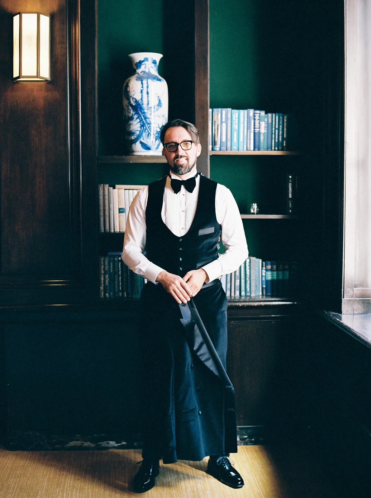 Groom standing in vest and bow tie in a library room with bookshelves and ceramic vase.