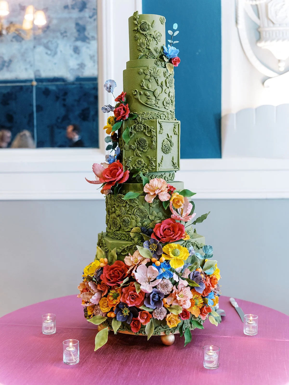 Tall green wedding cake with intricate floral detailing and colorful sugar flowers on reception table.