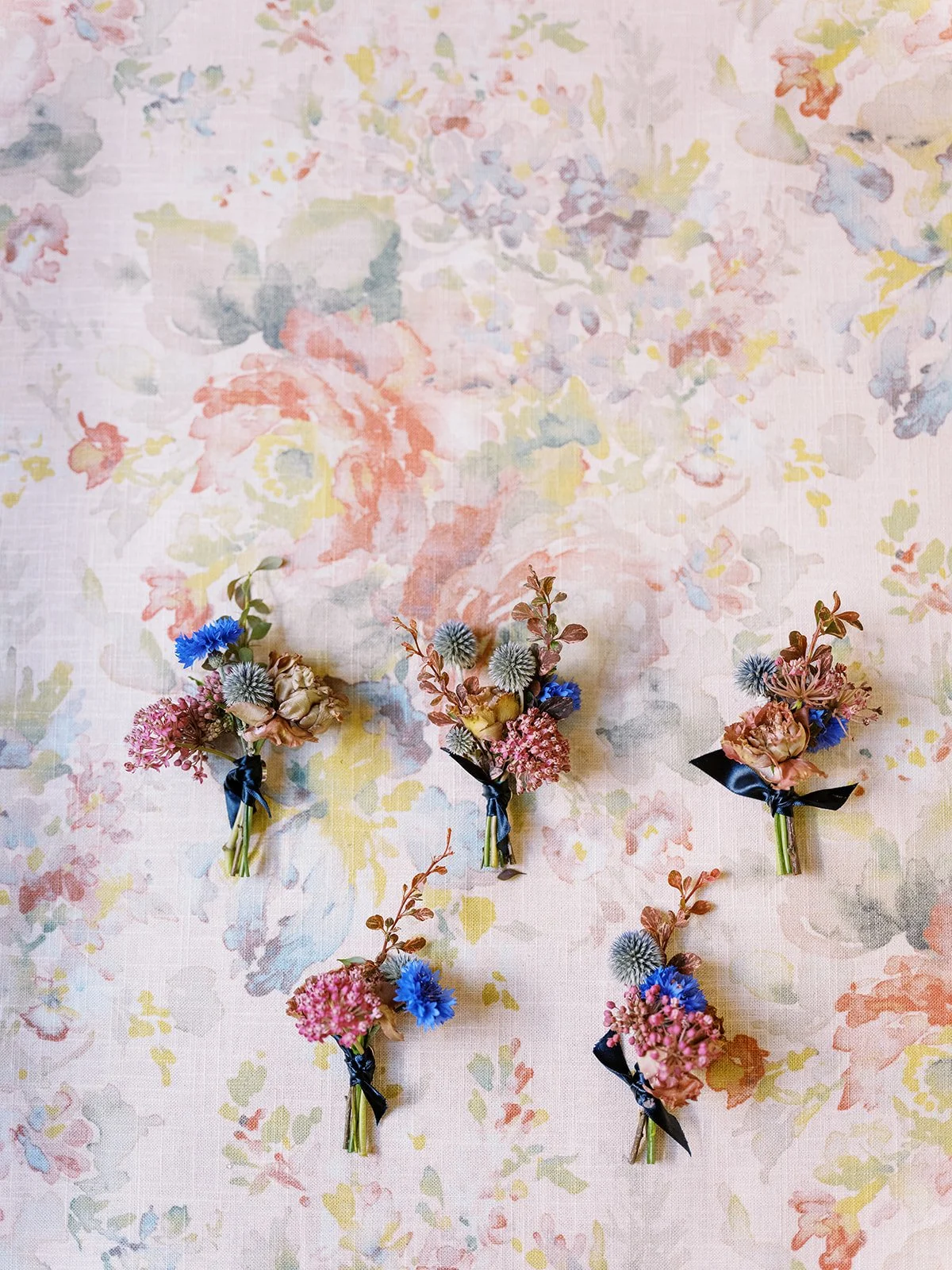 Five colorful boutonnières with blue thistle and pink flowers arranged on floral fabric background.