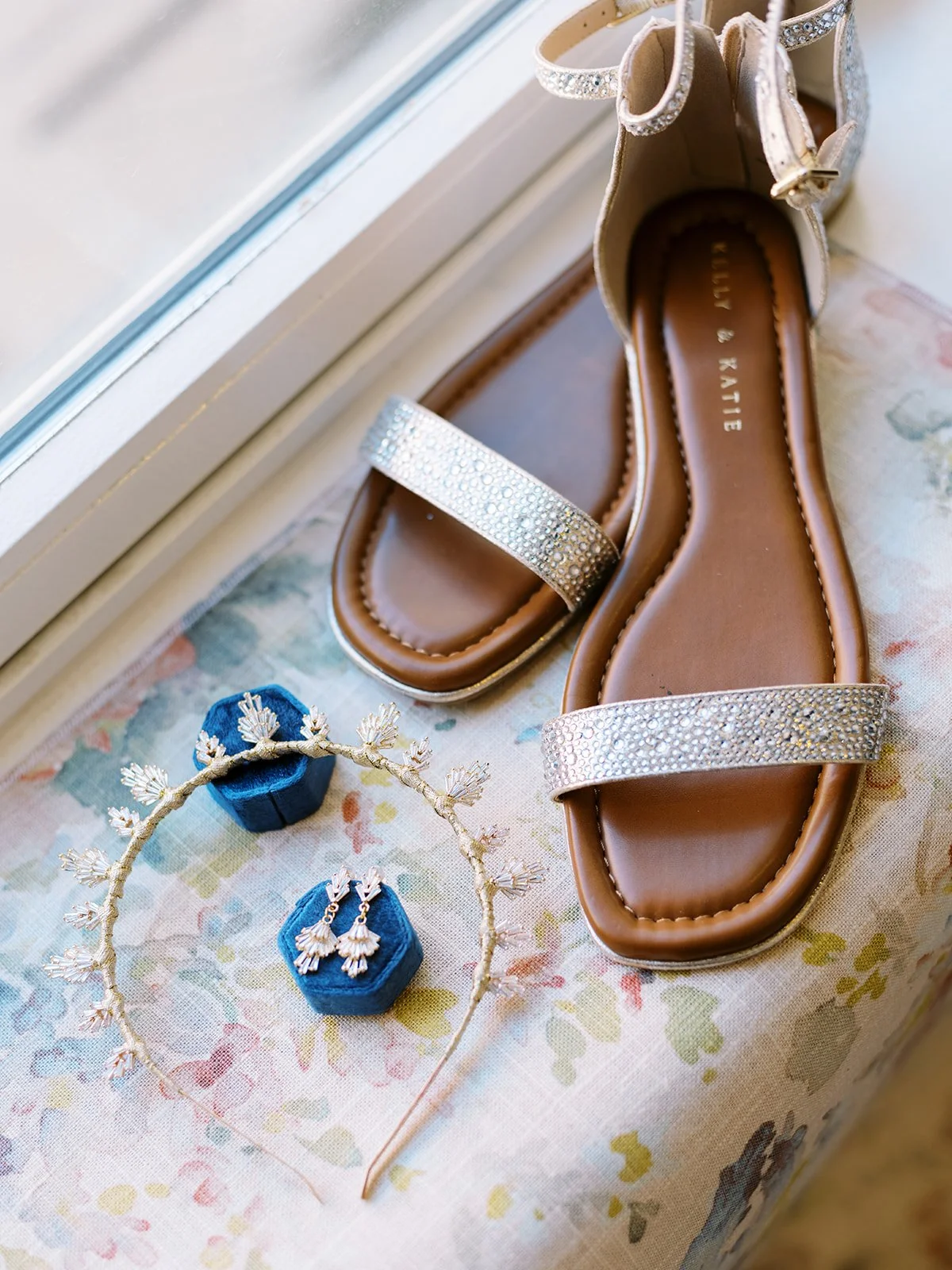 Bridal sandals, gold headpiece, and earrings arranged beside velvet ring boxes on floral fabric.