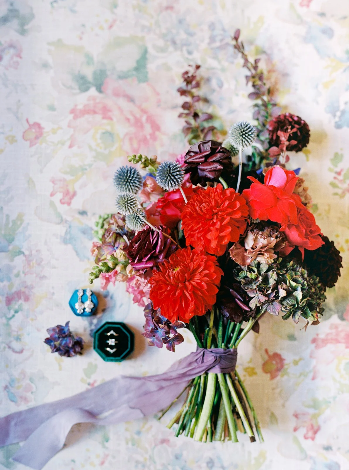 Colorful bridal bouquet with red, pink, and blue flowers beside wedding rings and earrings.