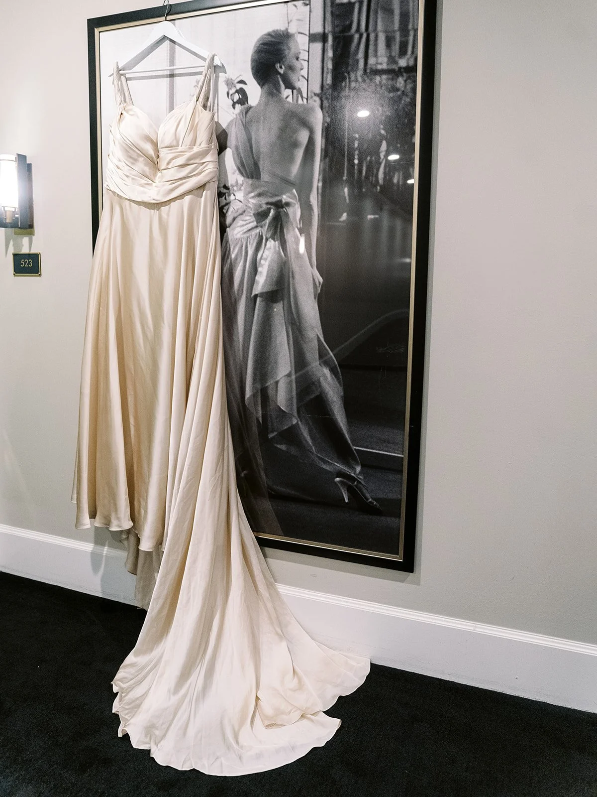 Silk wedding dress hanging from wall hook beside large framed black-and-white portrait.