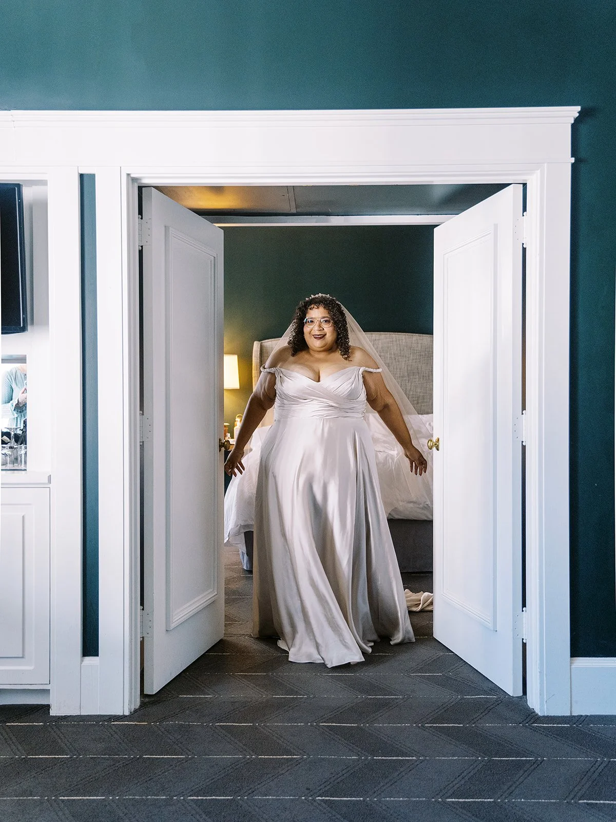 Bride in off-shoulder wedding gown standing in doorway inside hotel room before ceremony.