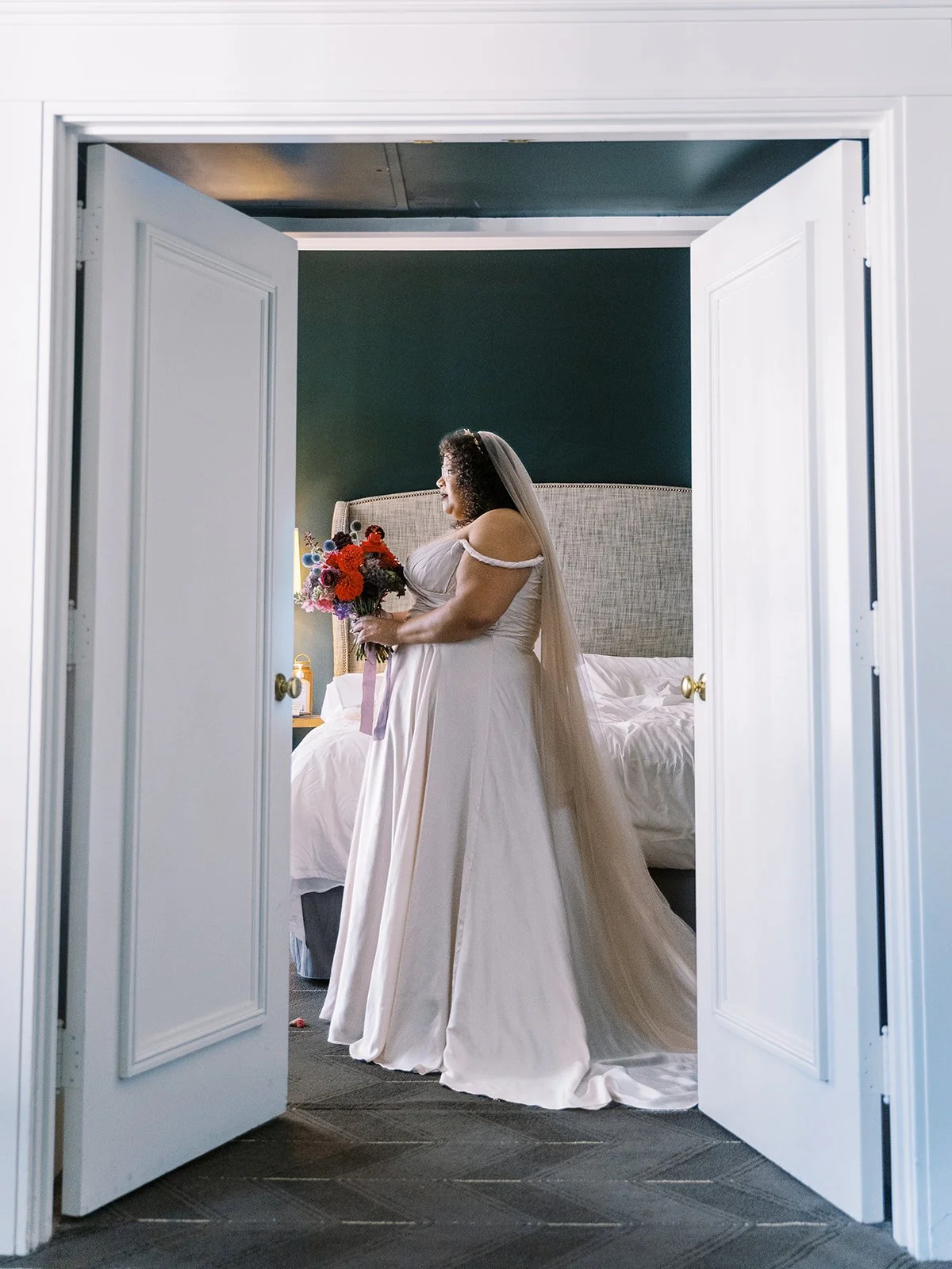 Bride in wedding gown holding colorful bouquet while standing in doorway of hotel suite.