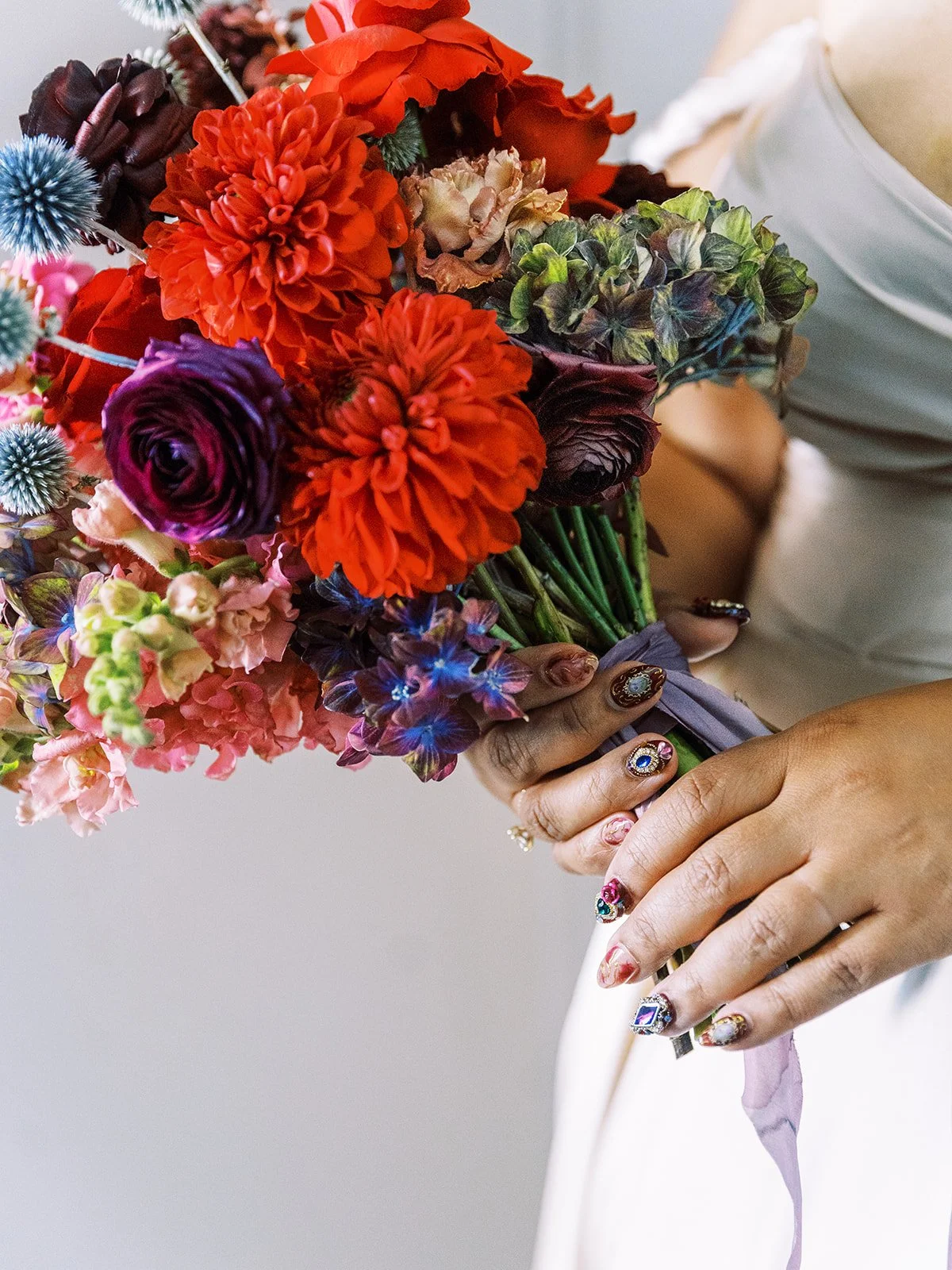 Close-up of bride holding vibrant bouquet with red dahlias and blue thistle flowers.