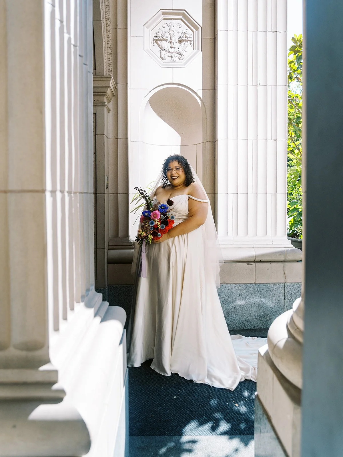 Bride in flowing gown holding colorful bouquet between tall stone columns.