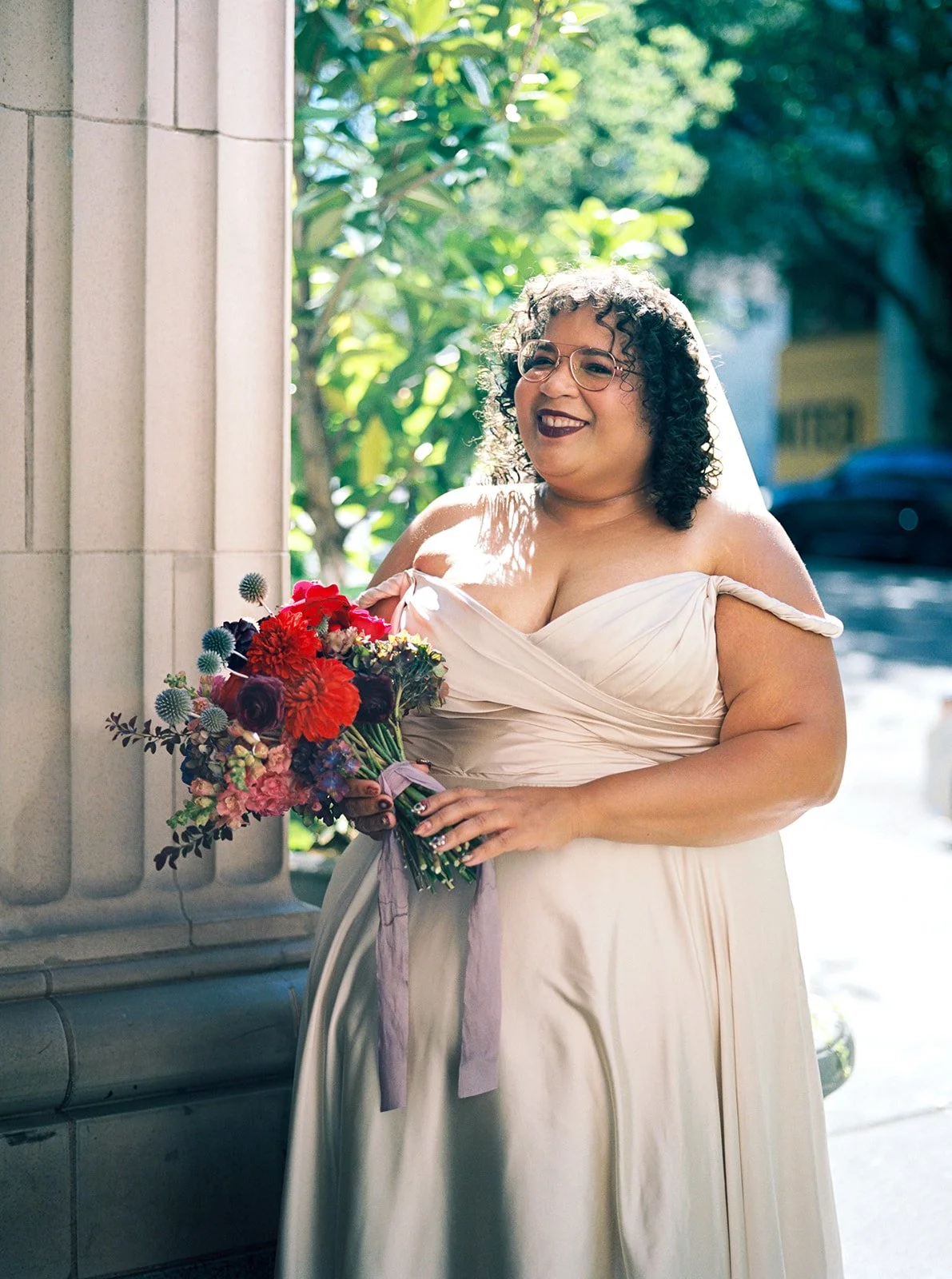 Bride holding bright bouquet beside stone column in soft outdoor light.