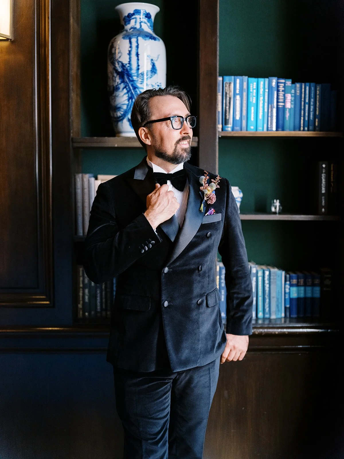 Groom in black tuxedo adjusting bow tie beside bookshelves in hotel library.