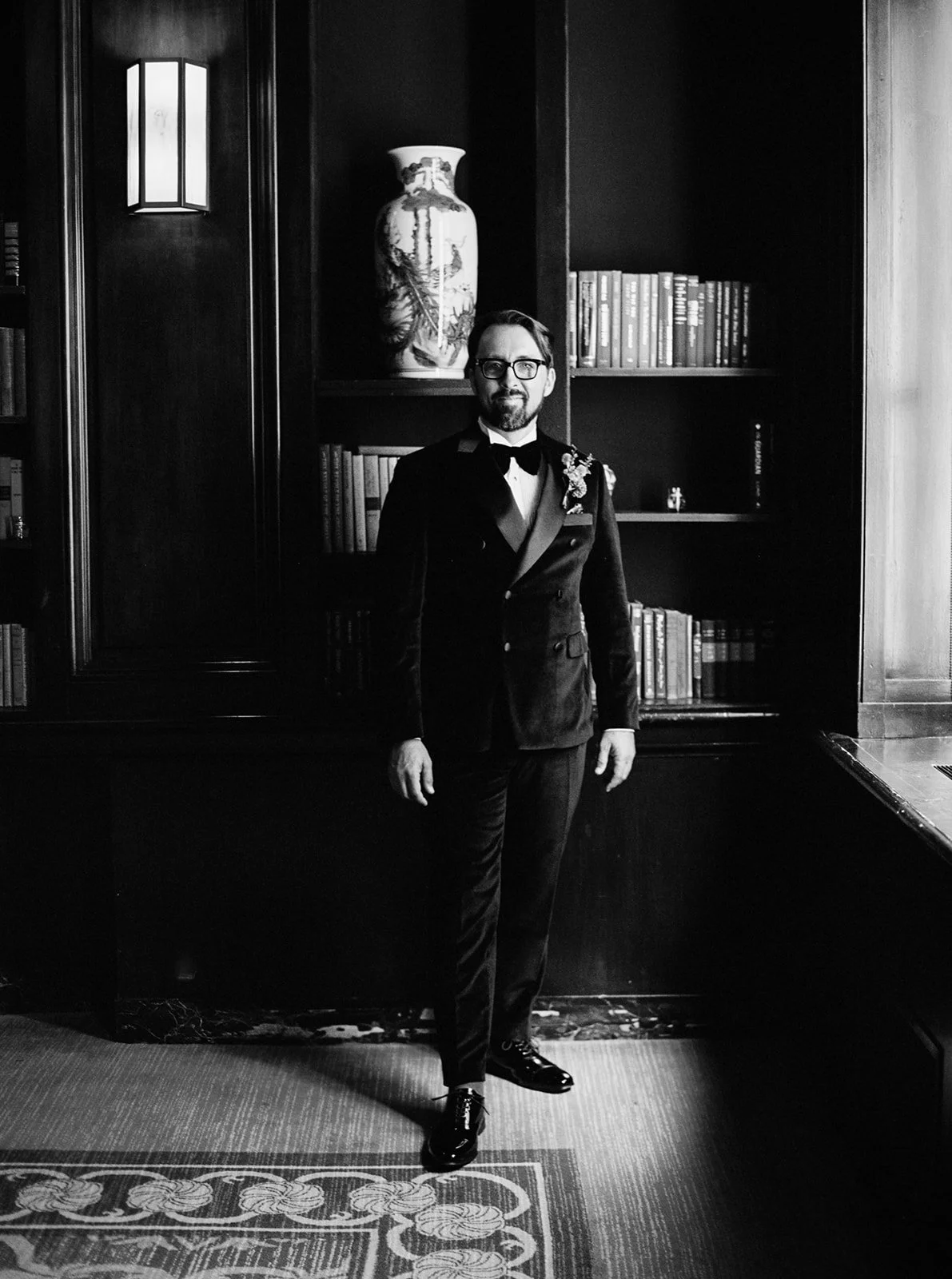 Groom in tuxedo standing in front of bookshelves in historic hotel library.