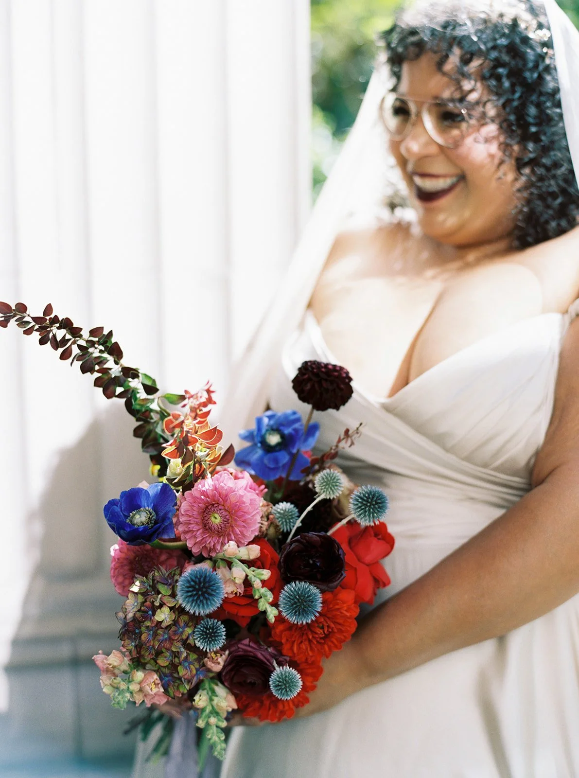 Bride laughing while holding colorful bouquet and wearing veil and glasses.