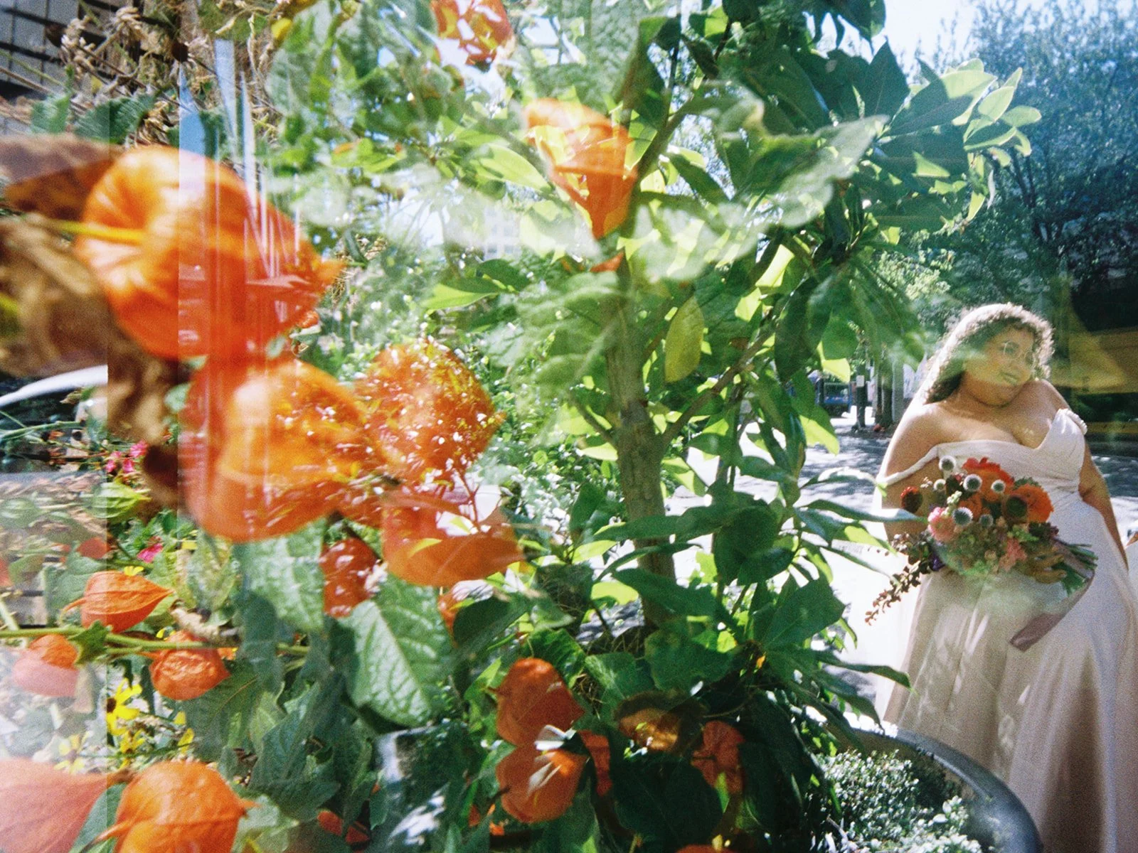 Bride holding colorful bouquet outdoors, framed through orange lantern plant leaves on sunny Portland street.