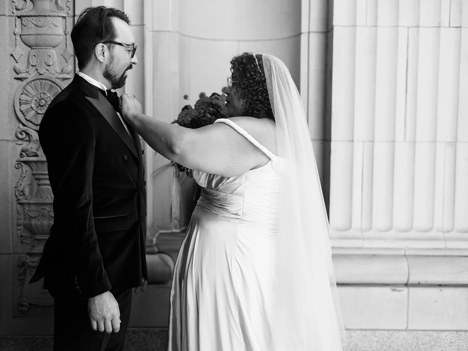 Bride adjusting groom’s bow tie during first look portrait beside stone columns.