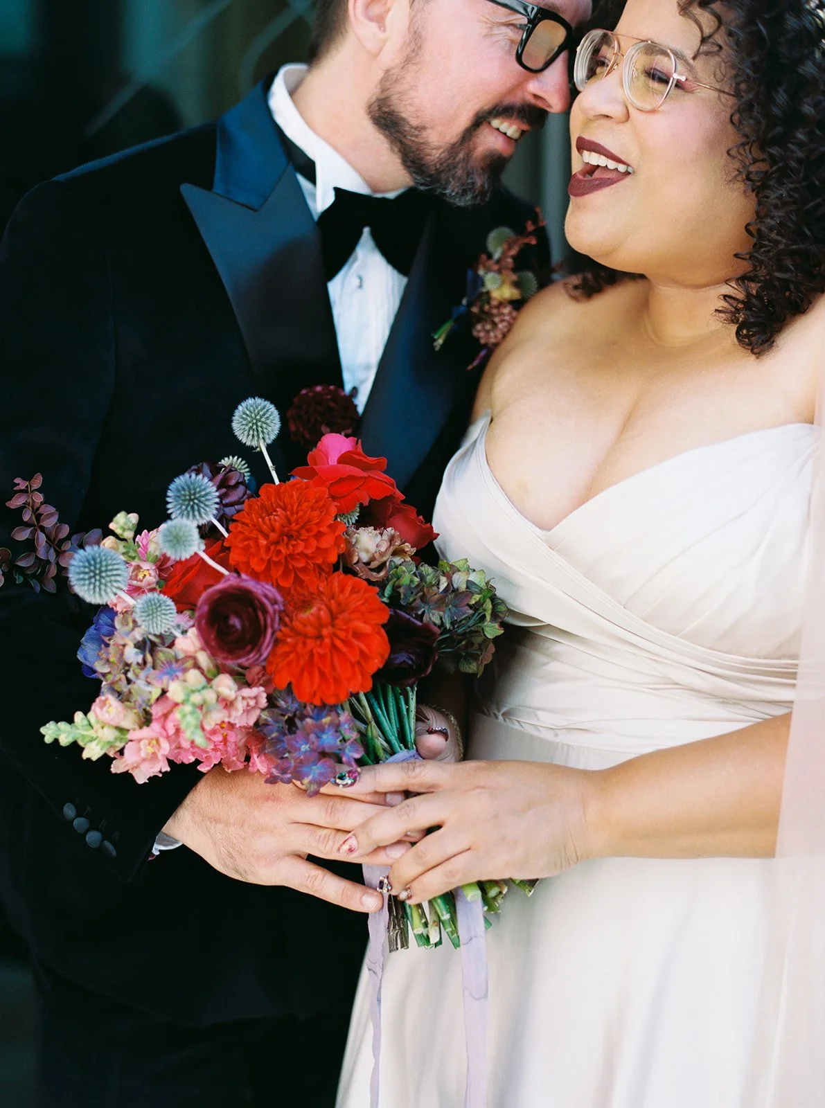 Close-up of bride and groom embracing with vibrant red and blue wedding bouquet.