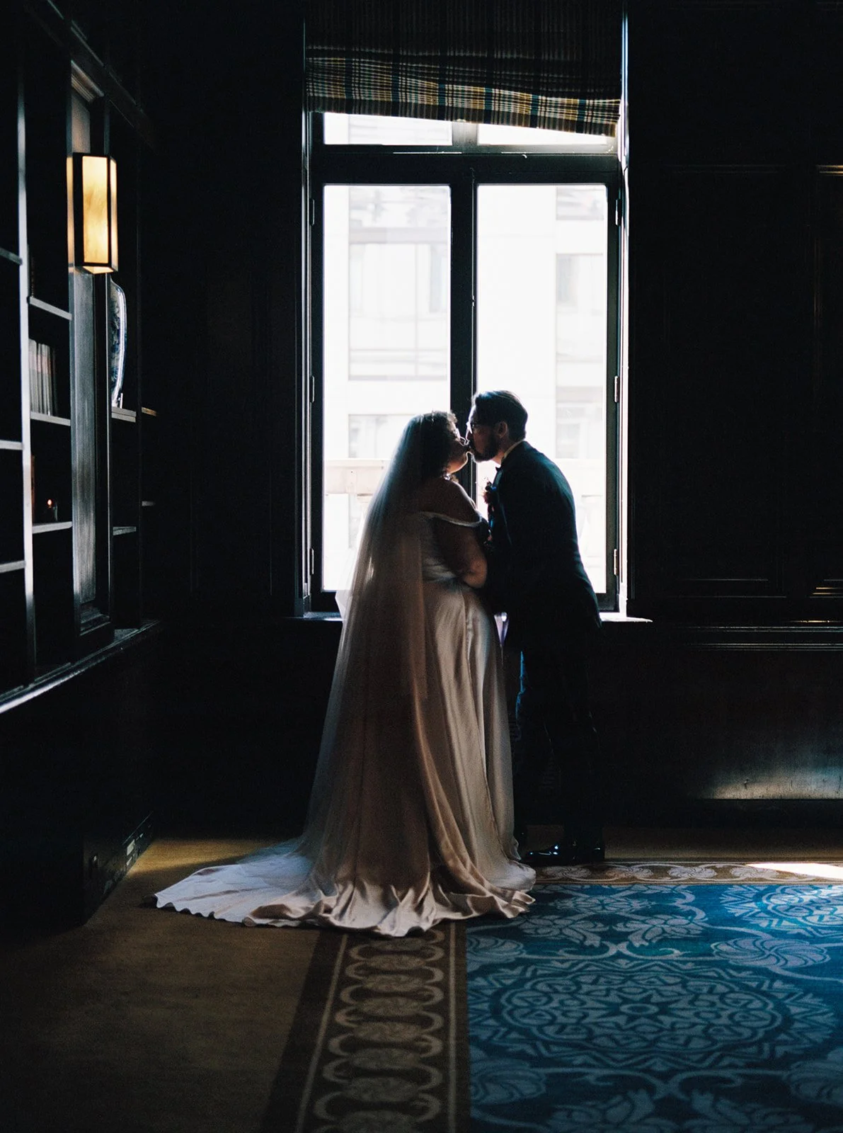 Bride and groom kissing in silhouette in front of bright window inside historic hotel library.