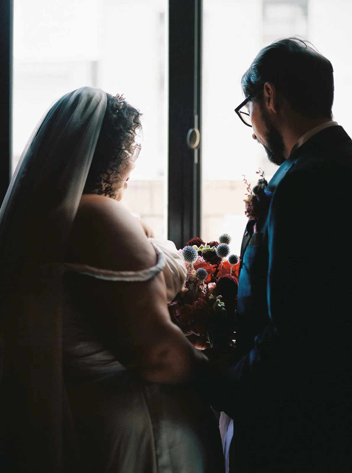 Bride and groom standing together by window holding colorful bouquet in dimly lit library.