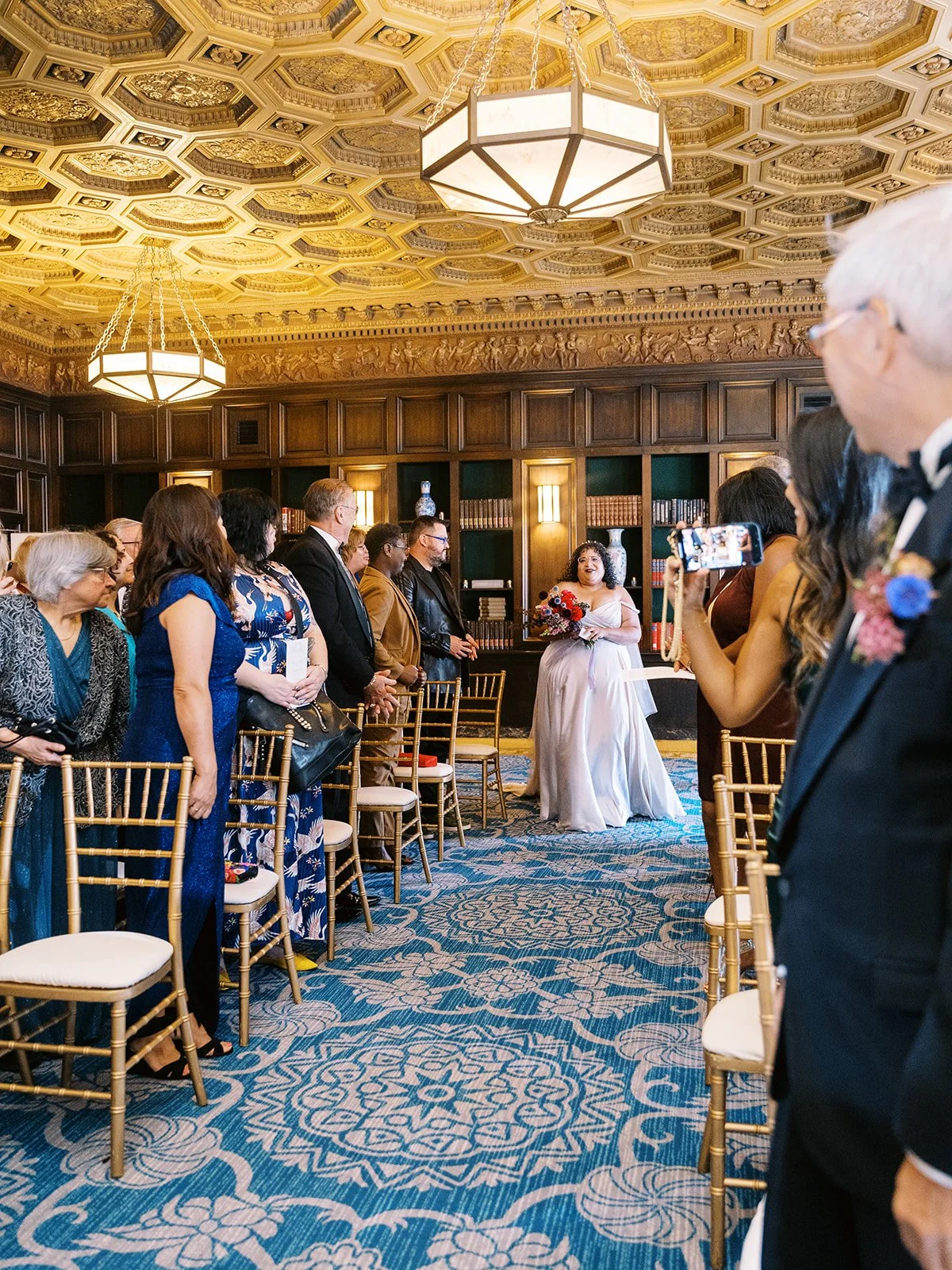 Bride walking down aisle holding colorful bouquet during indoor wedding ceremony.