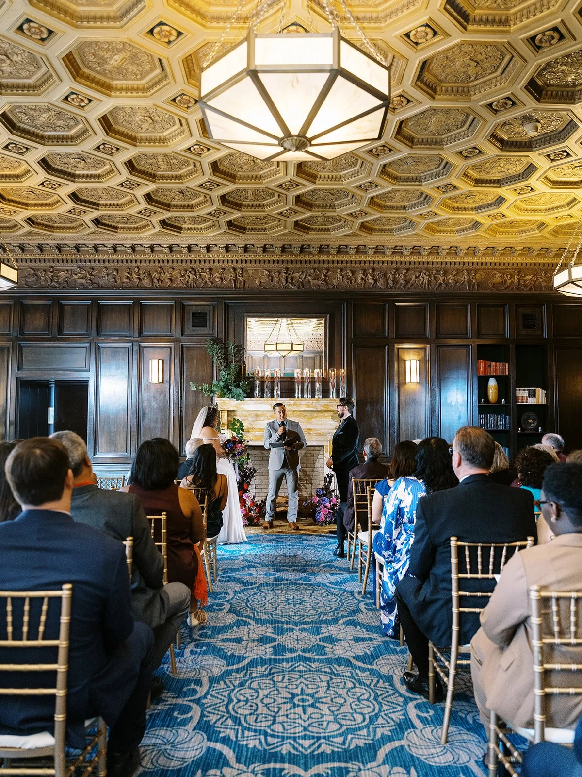 Wide view of indoor wedding ceremony in ornate wood-paneled library with gold coffered ceiling and marble fireplace.