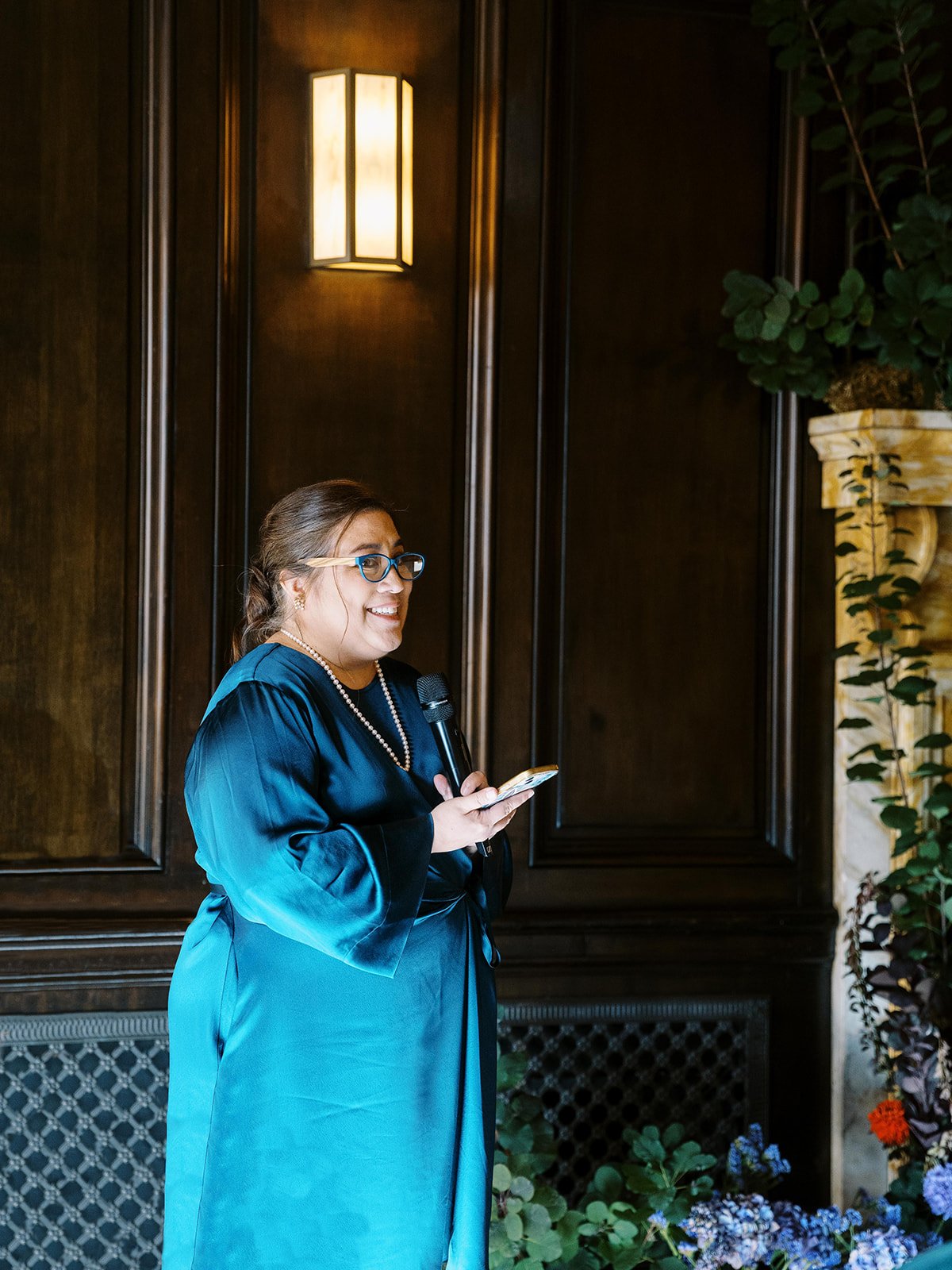 Wedding guest giving speech during ceremony beside fireplace with colorful floral installations.