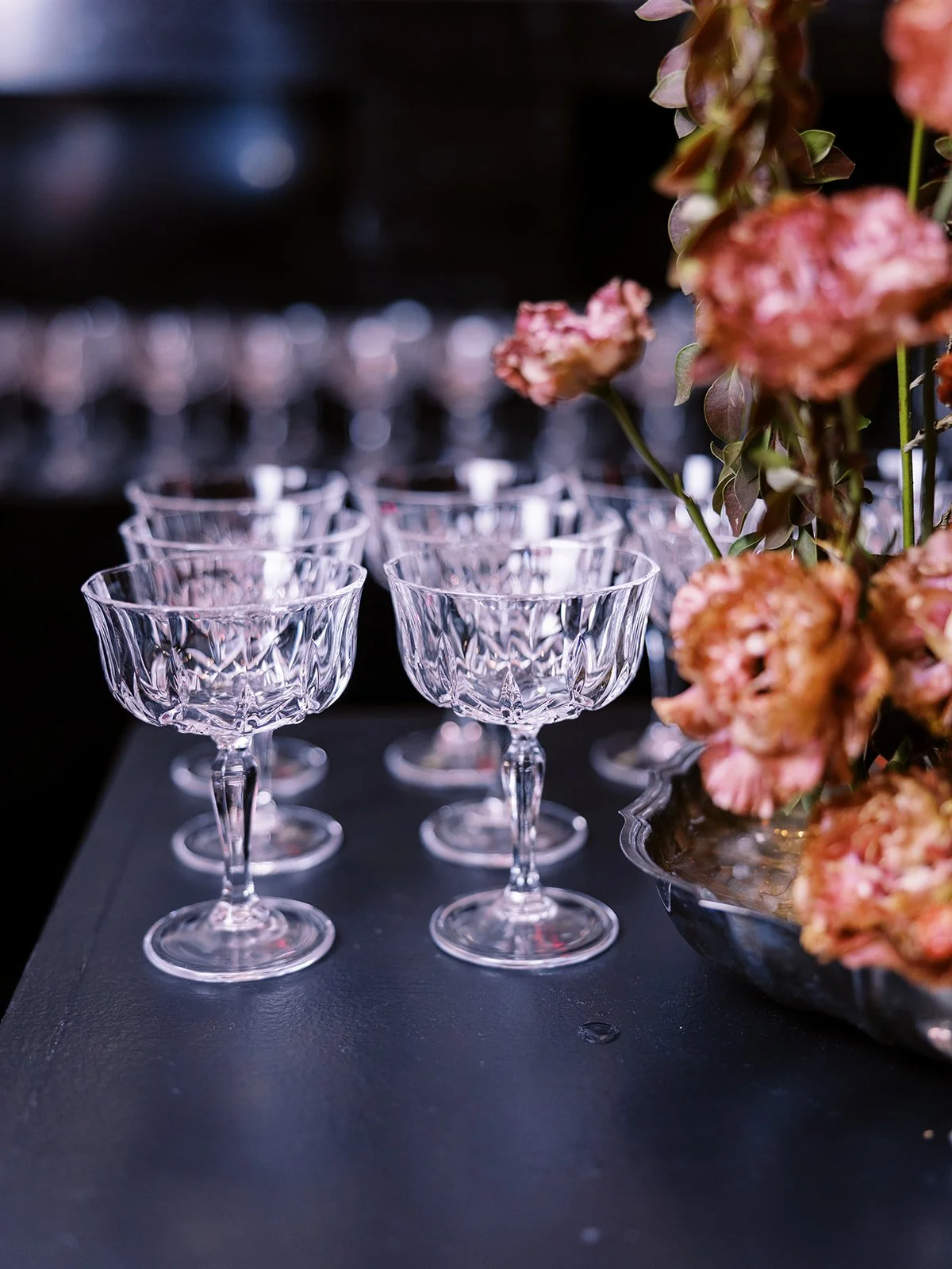 Crystal coupe glasses arranged on bar table beside floral arrangement for cocktail hour.