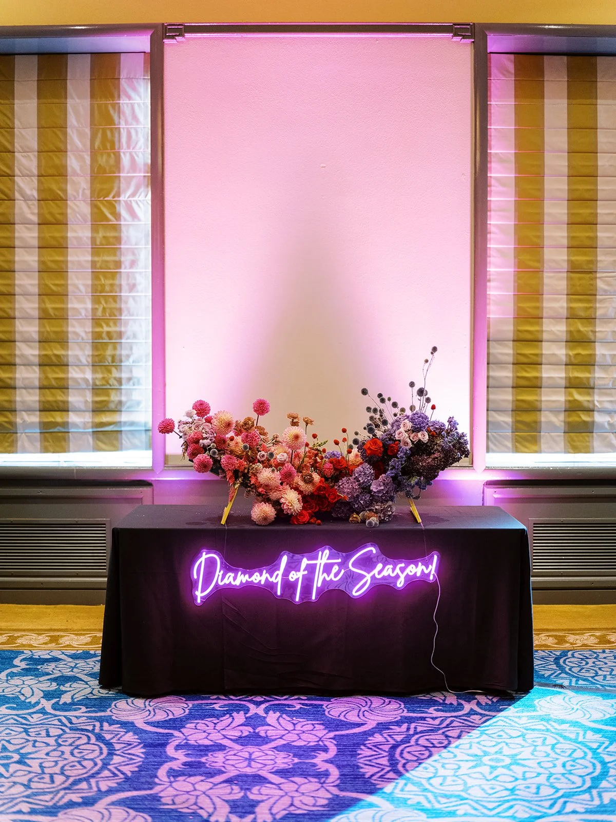 Long reception floral installation with pink, red, and purple flowers on table with neon “Diamond of the Season” sign.