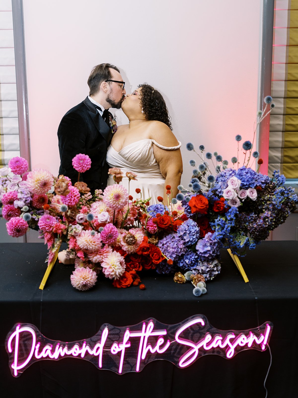 Bride and groom kissing behind vibrant floral arrangement and neon “Diamond of the Season” sign.