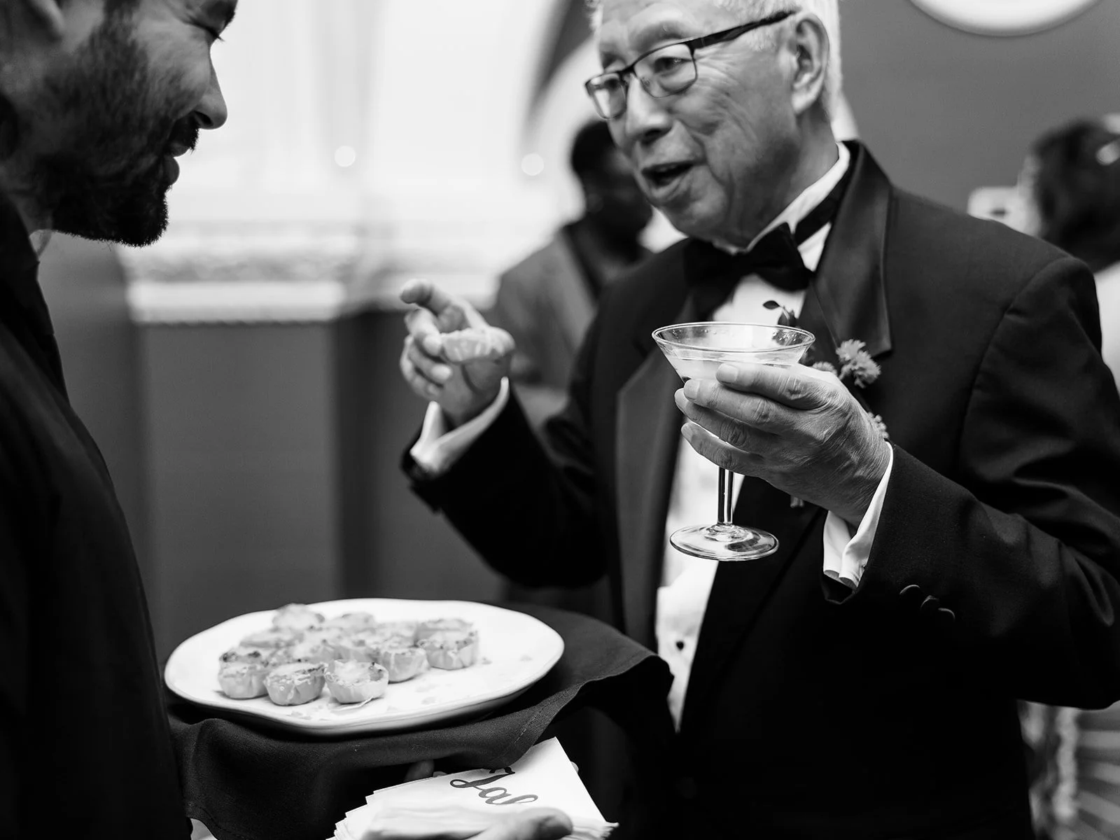 Wedding guest in tuxedo holding martini glass while chatting during cocktail hour.