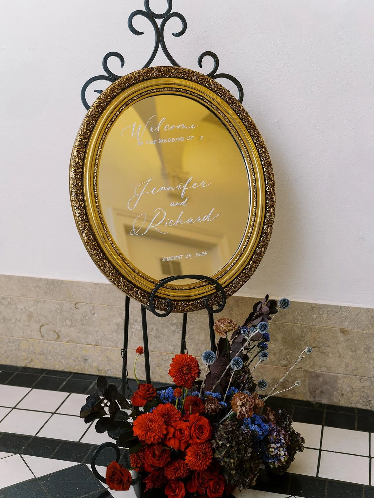 Gold framed mirror welcome sign reading “Jennifer and Richard” with red and blue floral arrangement below.
