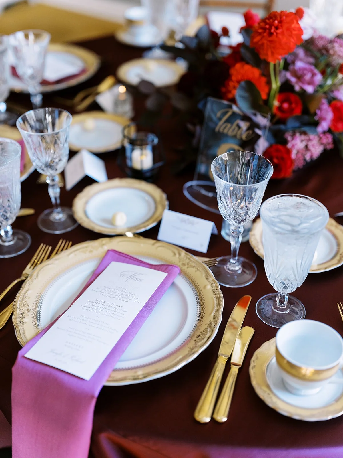 Reception place setting with gold flatware, pink napkin menu, and red floral centerpiece.