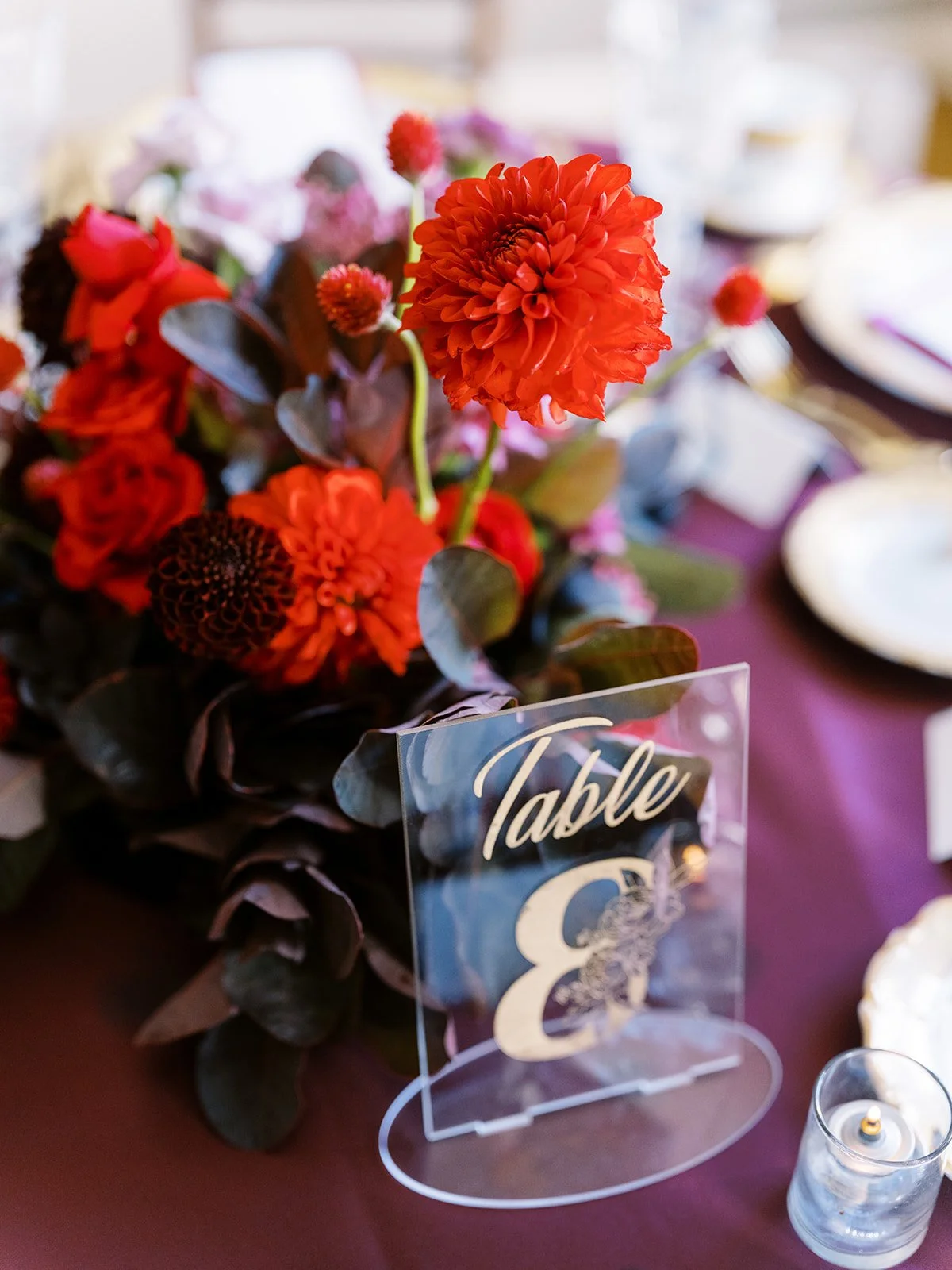 Red dahlia centerpiece with dark foliage beside acrylic “Table 8” sign on burgundy reception table.