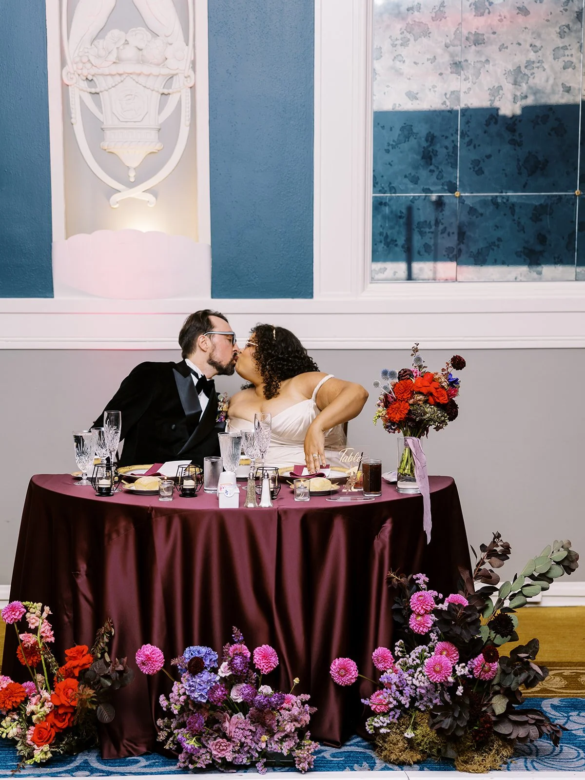 Bride and groom kissing at sweetheart table with burgundy linens and colorful floral arrangements.