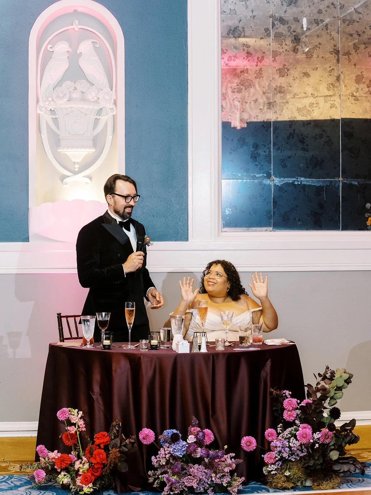Groom giving reception speech while bride smiles beside him at sweetheart table.