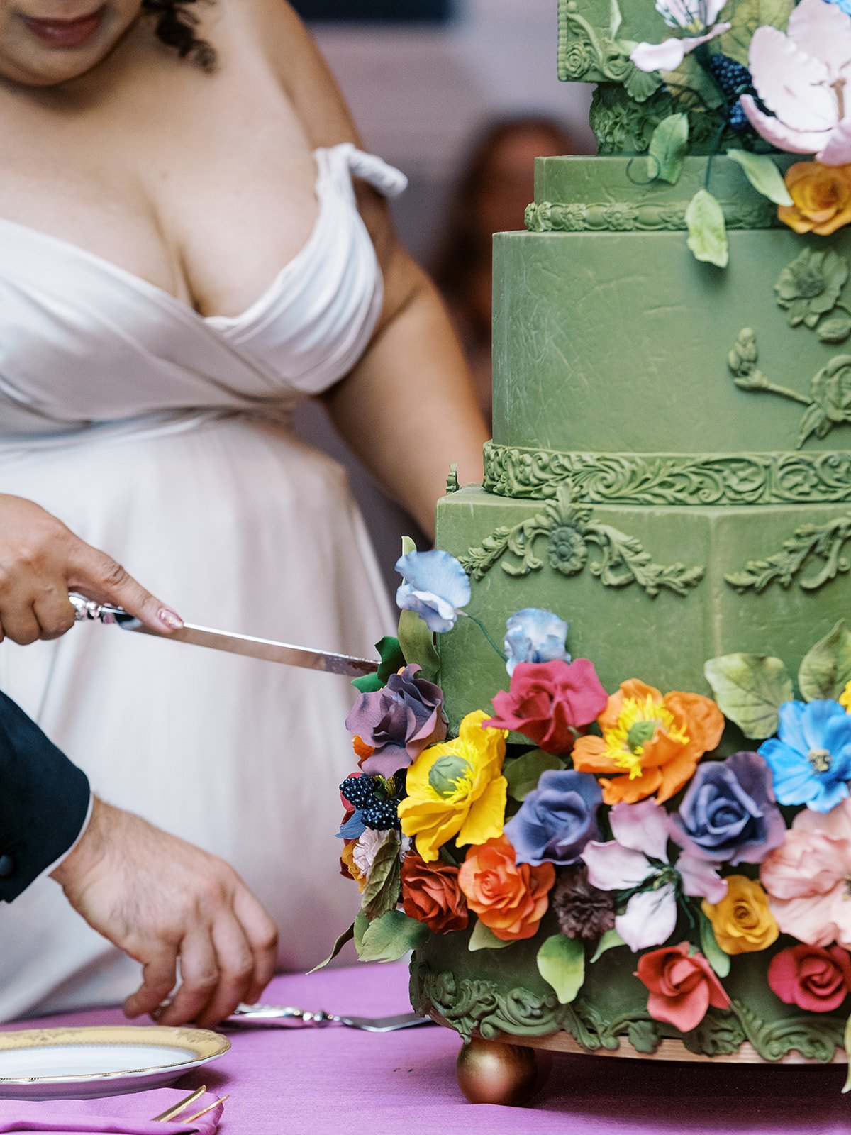 Bride and groom cutting ornate green wedding cake decorated with colorful sugar flowers.