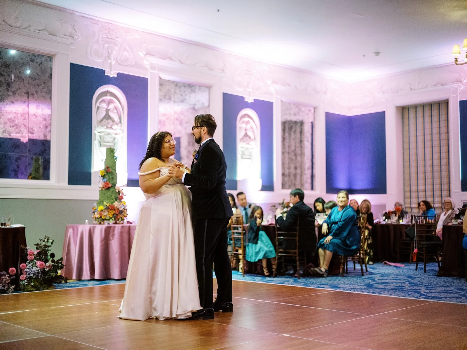 Bride and groom sharing their first dance in an ornate ballroom with purple uplighting, colorful wedding cake visible in the background