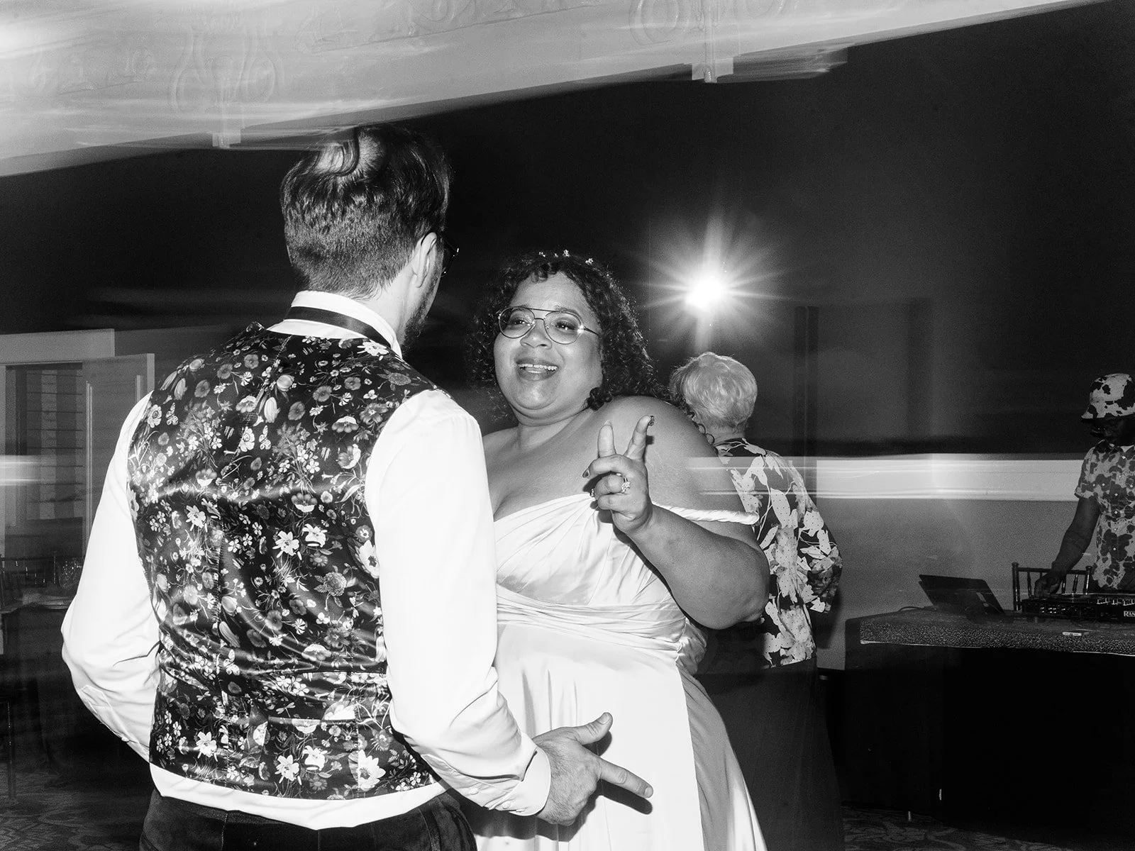 Black and white photo of the bride flashing a peace sign and grinning while dancing with the groom in his floral vest