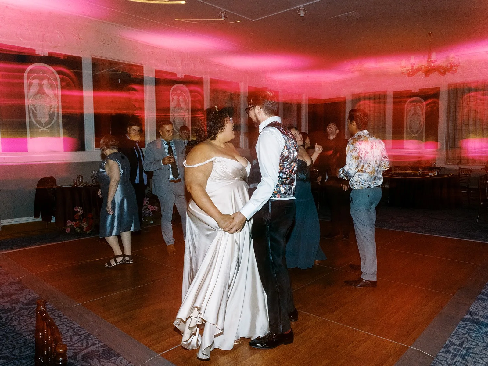Bride and groom dancing together under vivid pink and red uplighting, guests dancing around them in the ballroom