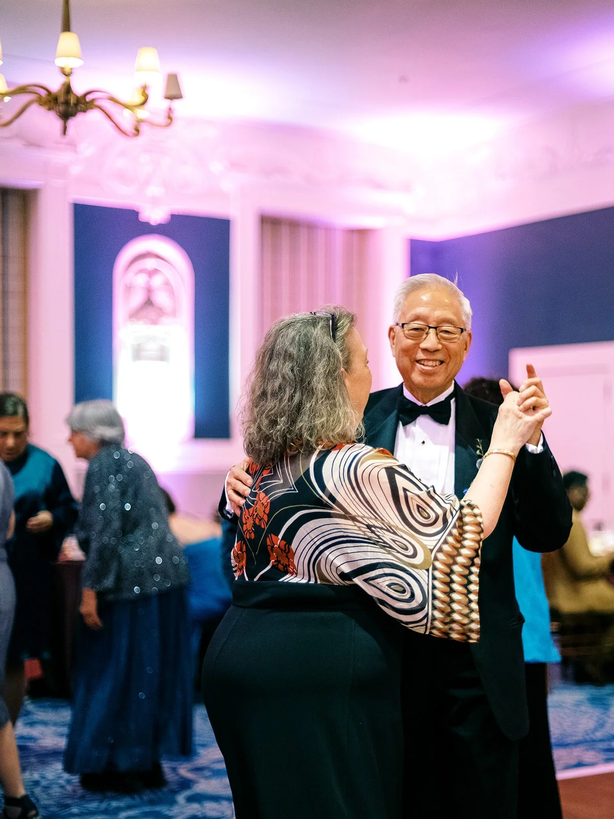 An older couple dancing joyfully together under warm pink uplighting in an elegant ballroom