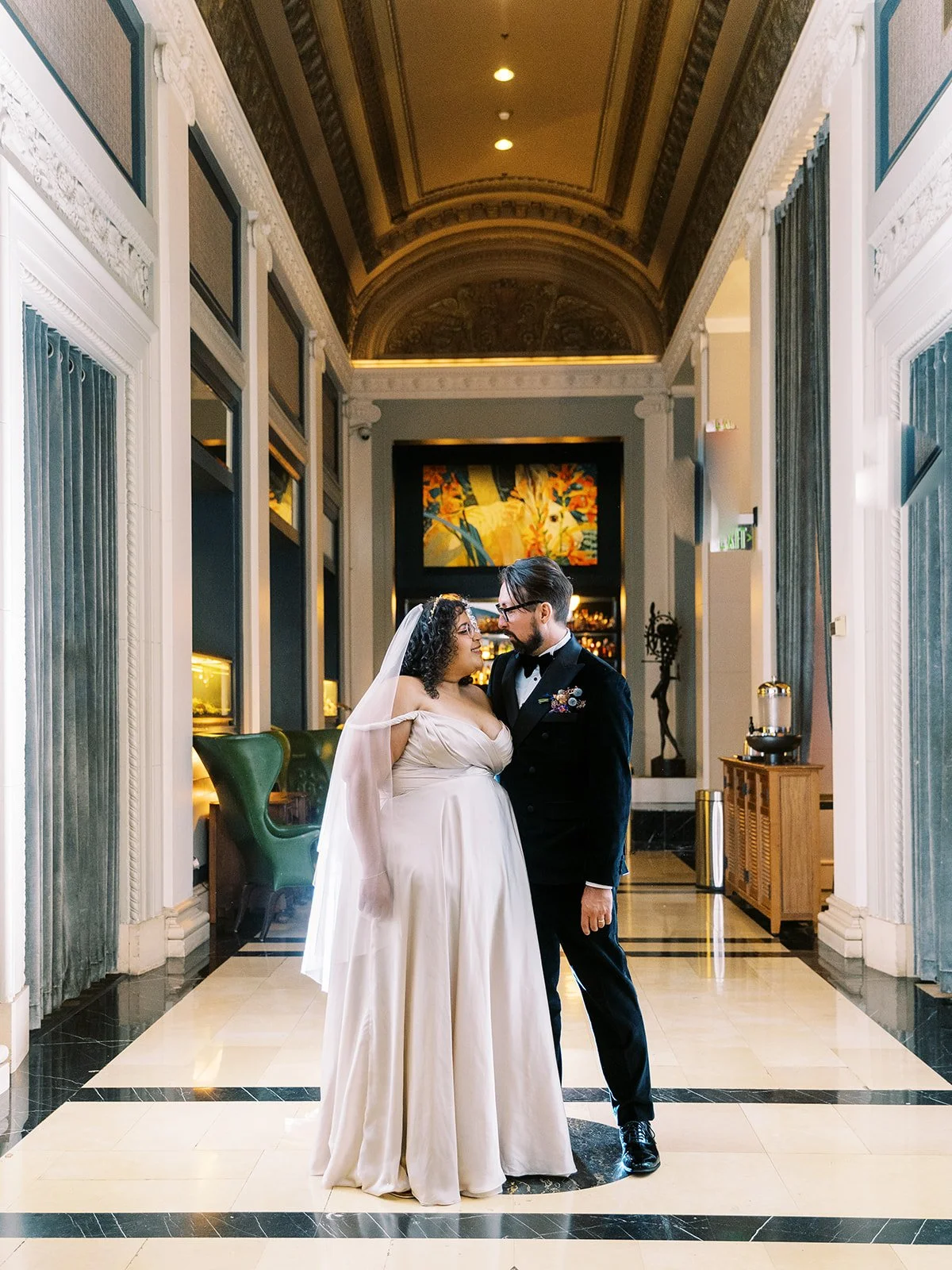 Bride and groom facing each other in an ornate hotel corridor with vaulted ceilings, marble floors, and a colorful painting in the background