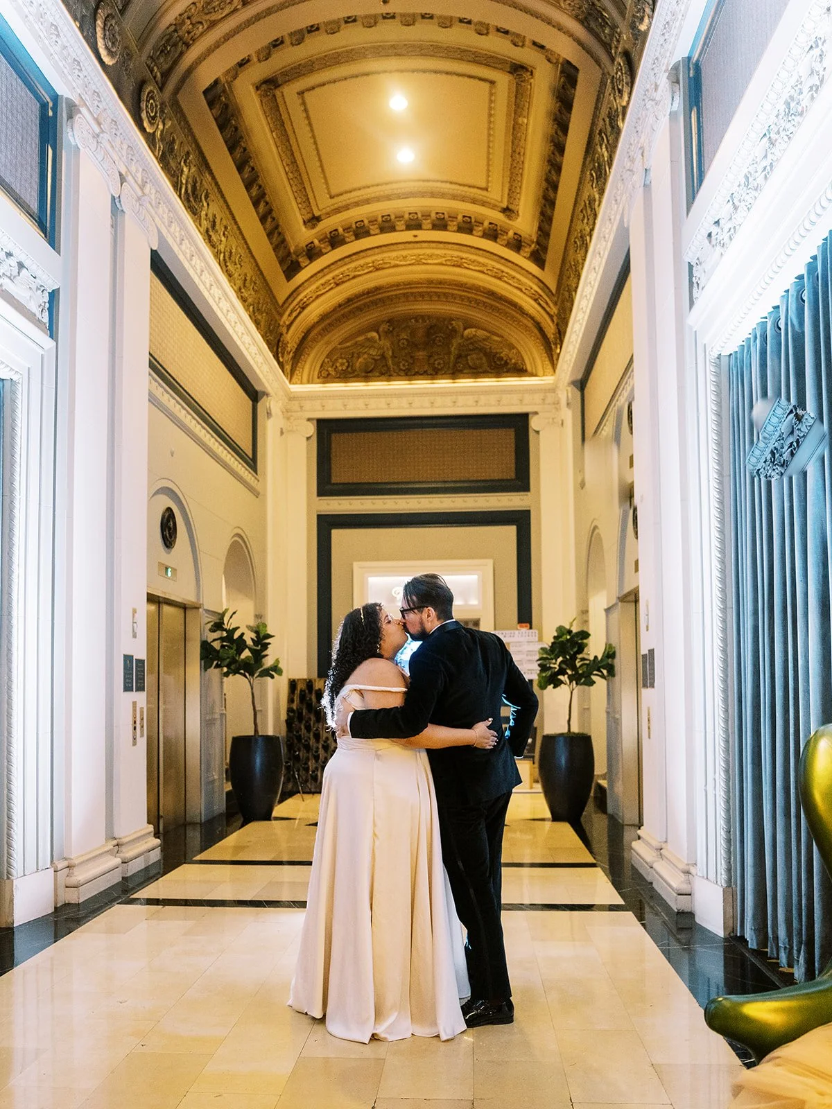 Bride and groom kissing in a grand hotel lobby with an ornate gilded barrel-vaulted ceiling and marble floors